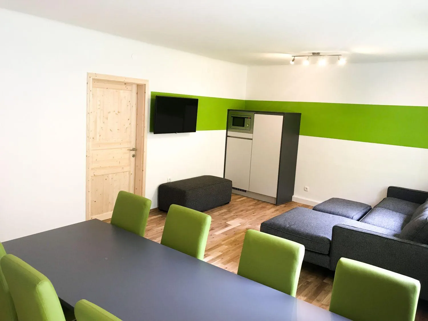 Gemeinschaftsraum mit großem Tisch, grünen Stühlen und TV an grüner Wand