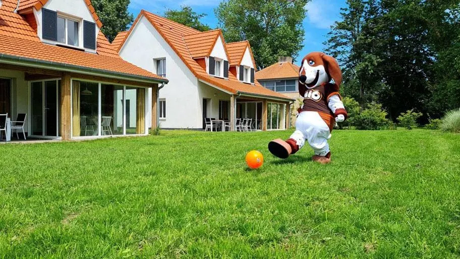 Maskottchen spielt Fußball auf Rasen vor typischen Ferienhäusern