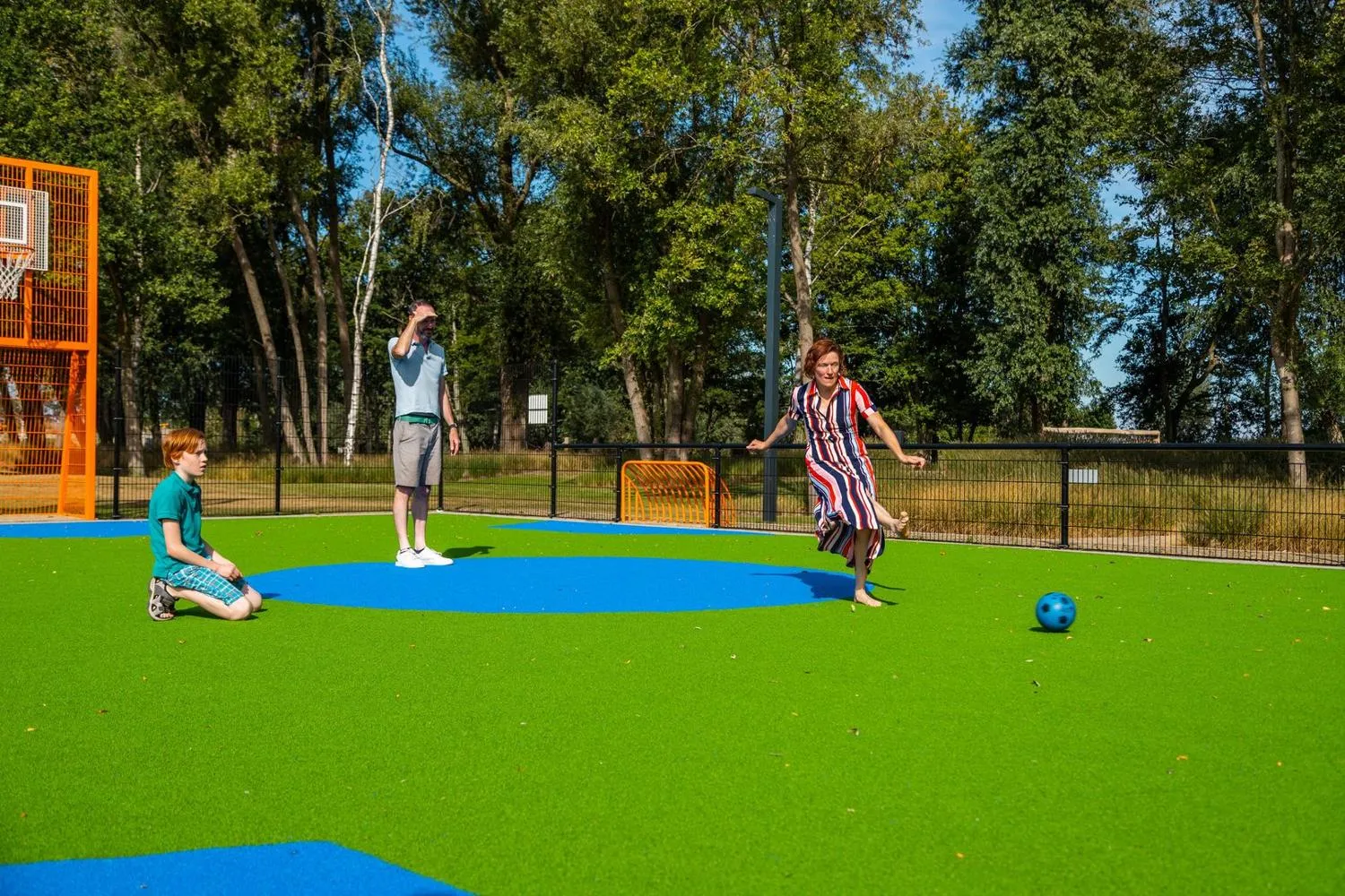 Spielfeld mit grün-blauem Kunstrasen unter Bäumen, Personen spielen Ball