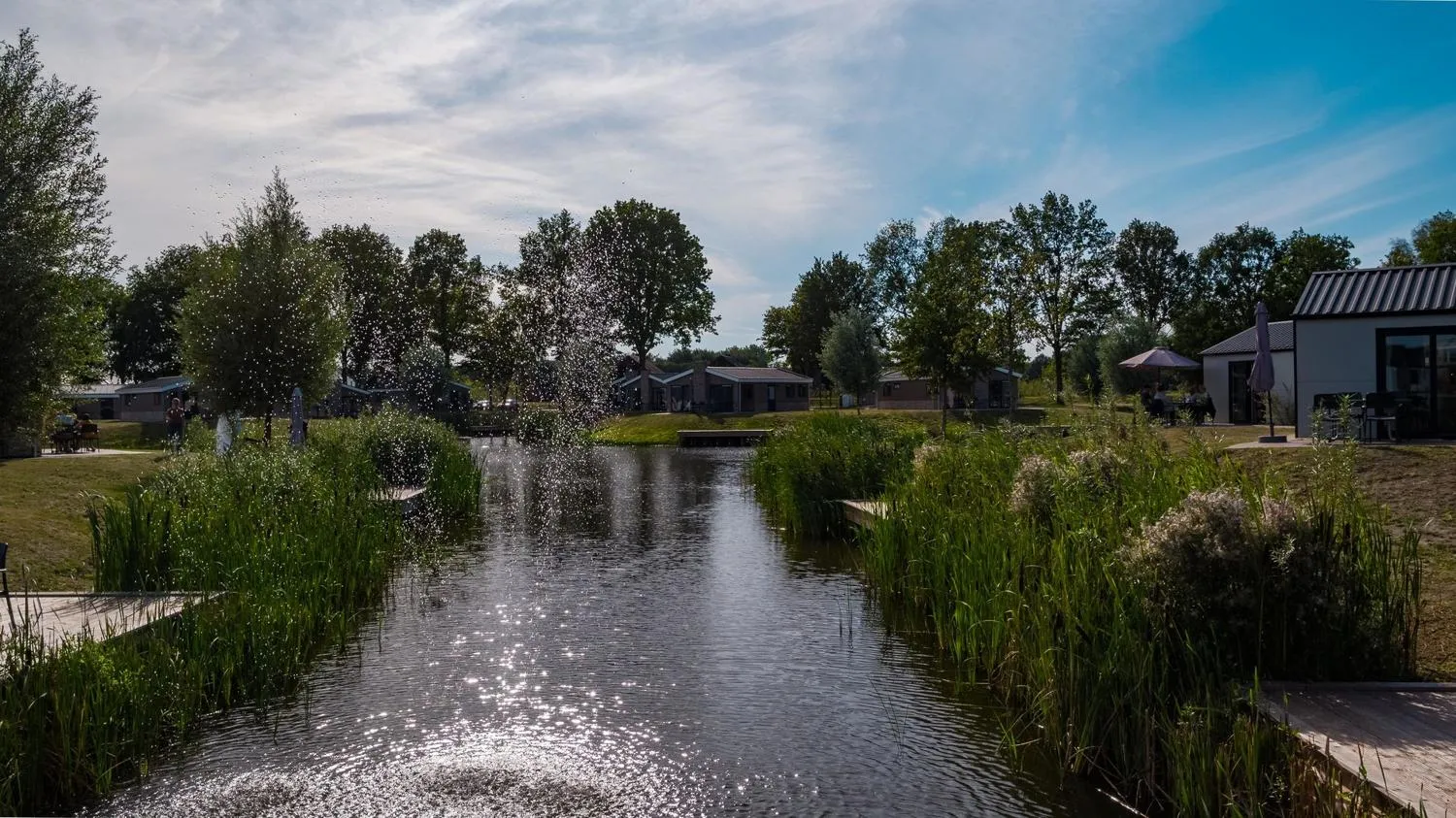 Wasserfontäne im Parkgelände, Ferienhäuser mit dunklen Dächern, grüne Uferzone